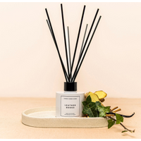 White bottle Leather Books reed diffuser with black sticks, set on a beige tray with green foliage accents.