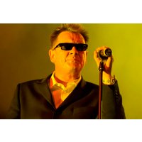 Man in black suit and sunglasses singing into a microphone on a yellow-lit stage.