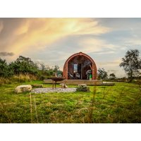 Rustic wooden pod cabin with arched roof, large glass doors, set in a grassy field with a fire pit and scenic sky.