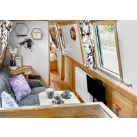 Narrowboat interior with wood paneling, floral curtains, grey sofa, purple cushion, and dining area with tableware.