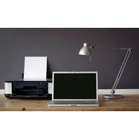 Laptop with blank screen, black printer with paper, and silver desk lamp on wooden table against gray wall.