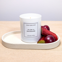 White Simon James Home soy wax candle labeled "Plum & Rhubarb," on a tray with fresh plums and rhubarb.