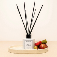 Plum & Rhubarb reed diffuser with black reeds in a white square bottle, placed on a beige tray with fruit accents.