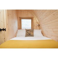 Wooden glamping pod interior with double bed, yellow blanket, and decorative pillow under a small window.