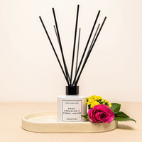 White diffuser bottle with black reeds, labeled "Rose, Geranium & Green Leaves," on tray with pink rose and yellow flowers.