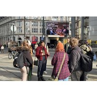 Group of people standing outdoors in a city, with backpacks and jackets, near a large digital billboard.