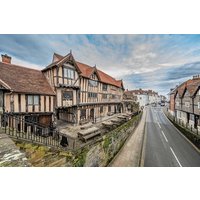 Historic timber-framed building with red-tiled roofs and gardens, adjacent to a narrow street, under a cloudy sky.