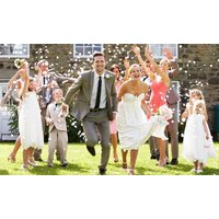 Bride and groom joyfully running with wedding party in colorful attire, surrounded by confetti on a sunny day.