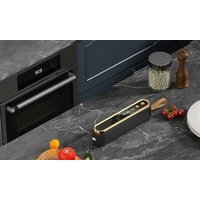 Black and gold rectangular knife sharpener on marble countertop near vegetables and spice jars.