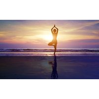 Silhouette of a person in tree pose on a beach at sunset, with vibrant purple and orange sky reflecting on water.