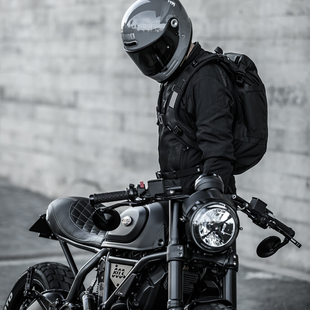 Black Kriega R16 waterproof backpack with adjustable straps, featuring a sleek design, mounted on a rider in motorcycle gear, next to a stylish motorcycle.