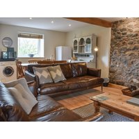 Cozy cottage living room with brown leather sofas, stone wall, wooden coffee table, and kitchen area with appliances.