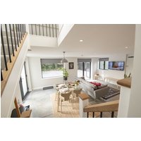 Modern living room with beige sofa, wooden dining table, large windows, and staircase with black railings.