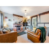 Cottage living room with brown sofas, brick fireplace, TV, and rustic wooden beams.