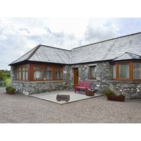 Stone cottage with slate roof, large windows, red bench on patio, and potted plants, under a cloudy sky.