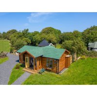 Wooden cottage with green roof, large windows, and small porch, surrounded by lush greenery and clear blue sky.