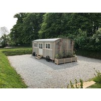 Wooden cottage with white-framed windows, metal roof, and wheelbase, set on gravel with surrounding greenery.