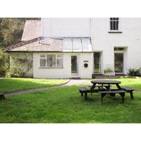 White cottage with glass conservatory, surrounded by lush green lawn and wooden picnic table in foreground.