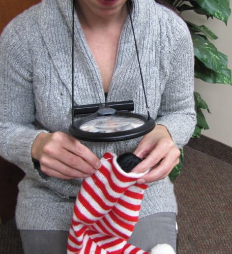 Hands-free lighted magnifier with a black frame and adjustable neck strap, featuring a large lens and a smaller 4x spot lens, used by a woman in a gray sweater while crafting.