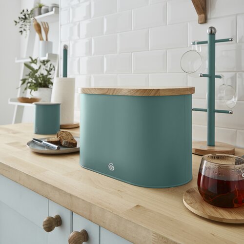 Teal Nordic oval bread bin with a wooden cutting board lid, placed on a light wood countertop, alongside a cup of tea and kitchen accessories.