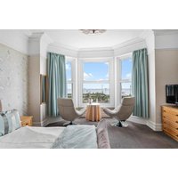 Hotel room with bay window, sea view, two modern chairs, small table, light blue curtains, and a flat-screen TV.