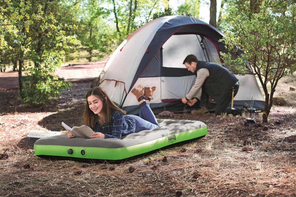Inflatable Bestway mattress in green and gray, featuring a soft velvety surface, set up outdoors with a woman reading and a man adjusting a tent in the background.