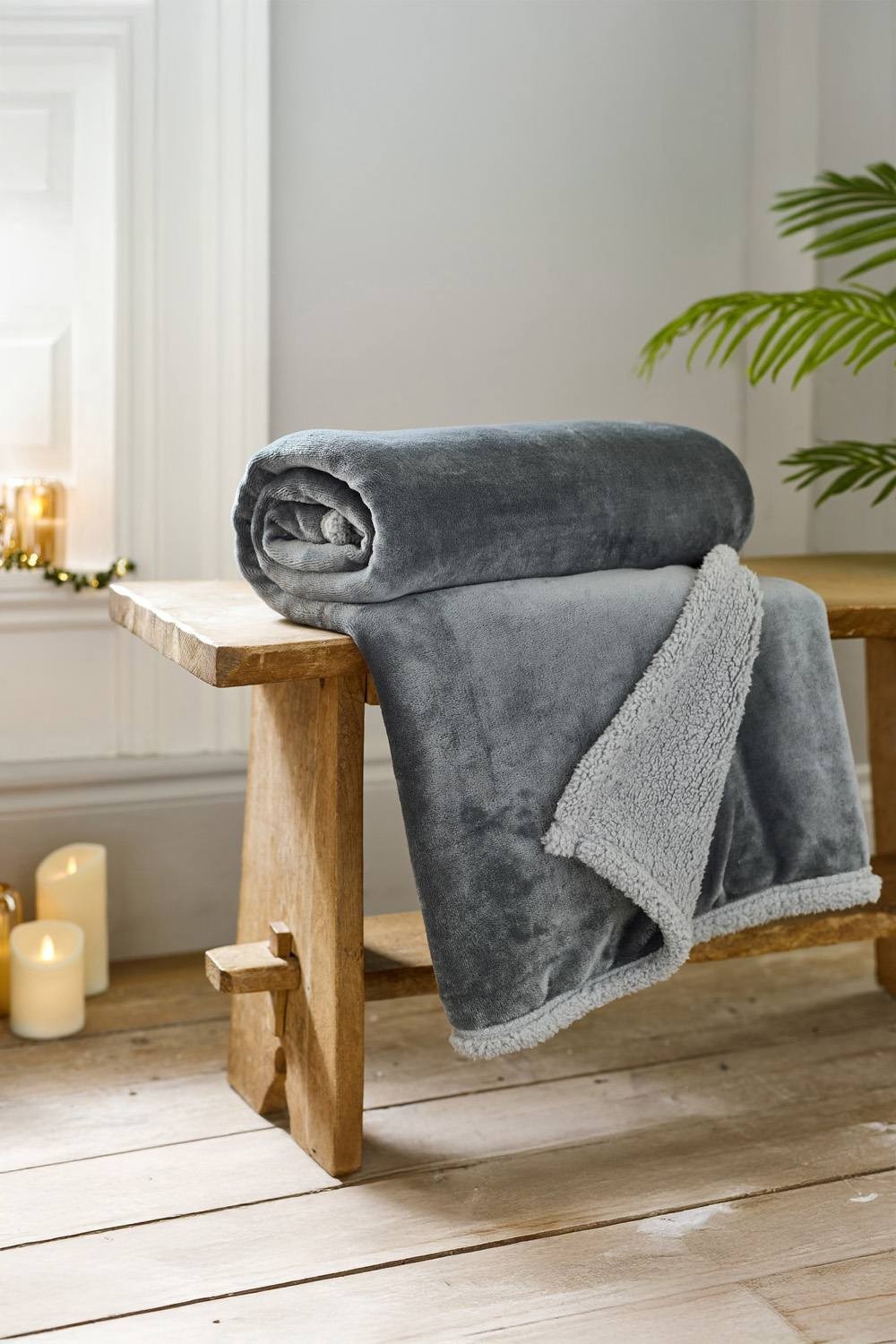 Super soft silver flannel and sherpa throw blanket, measuring 130 x 170 cm, neatly rolled and draped over a wooden bench, with candles and greenery in the background.