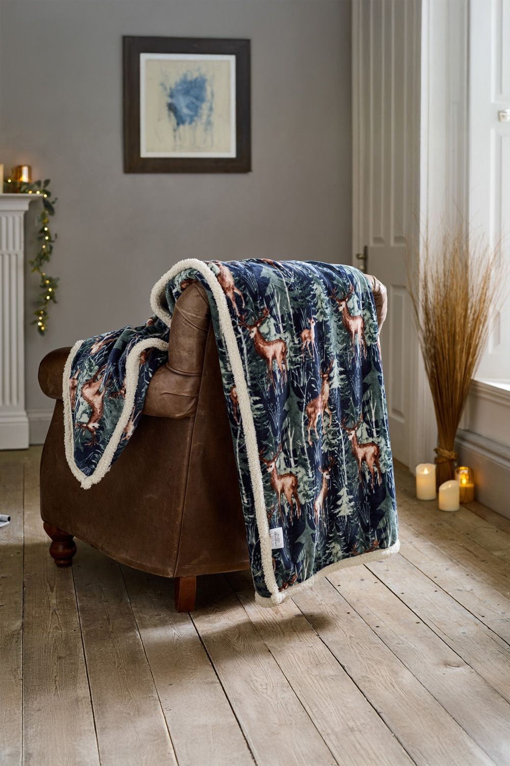 Navy forest print reversible fleece blanket featuring stag motifs, trimmed with soft cream sherpa, measuring 140x180cm, draped over a brown leather armchair.