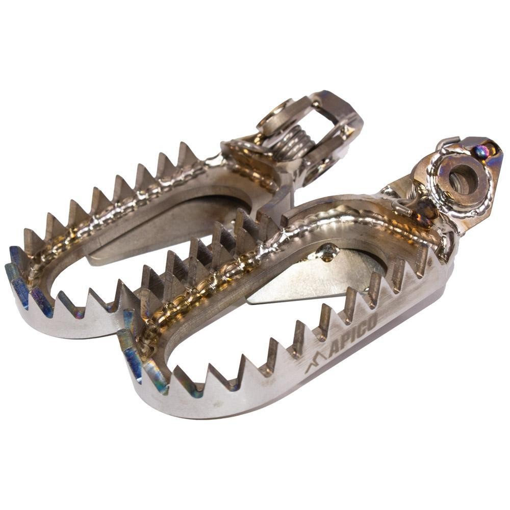 Open design titanium foot pegs with a polished finish, featuring serrated edges for grip, designed for various KTM models from 2023 to 2025, marked with the Apico logo.