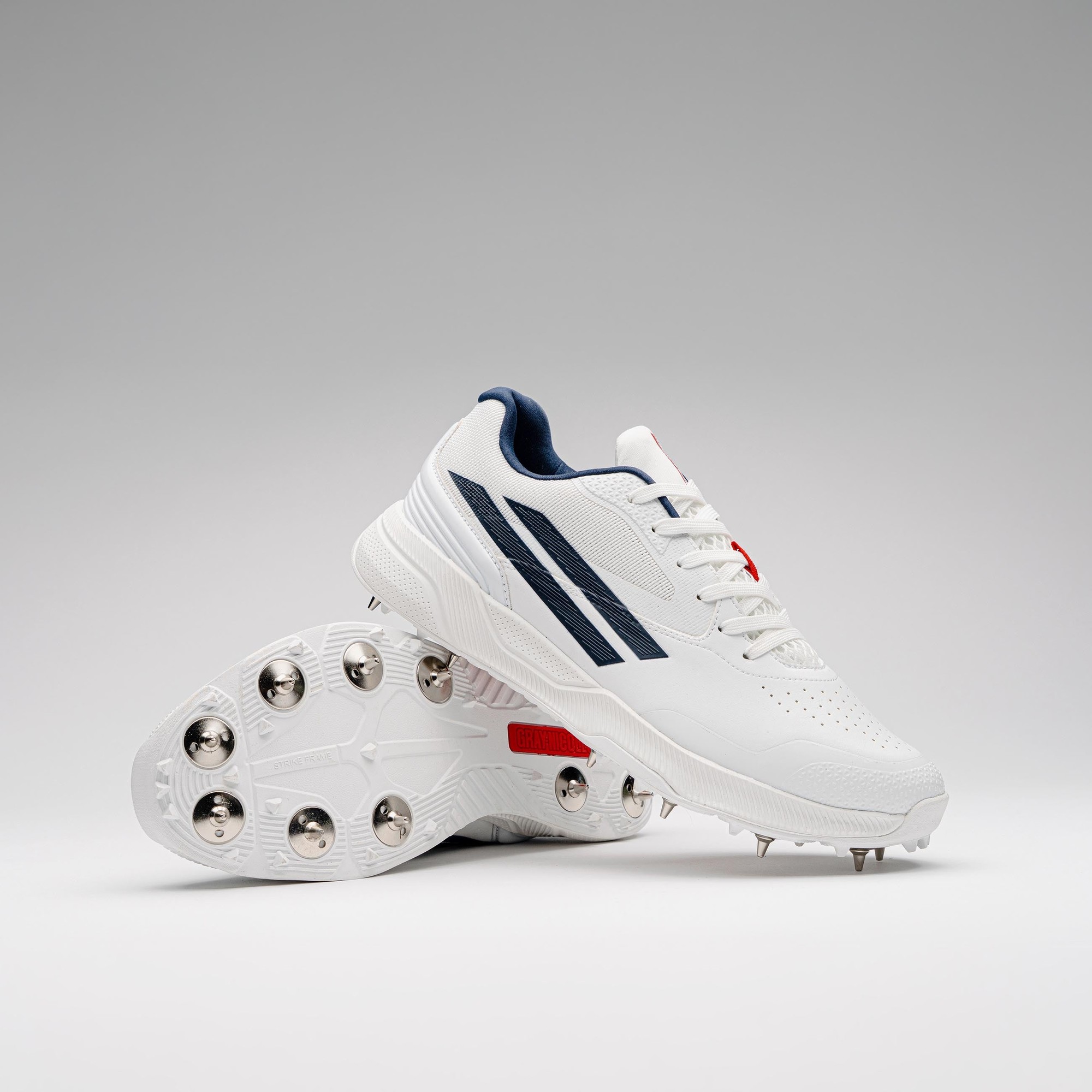 Gray-Nicolls Players 4.0 adult spike shoes in white with navy stripes, featuring a textured upper and metallic spikes on the sole for traction on the field.