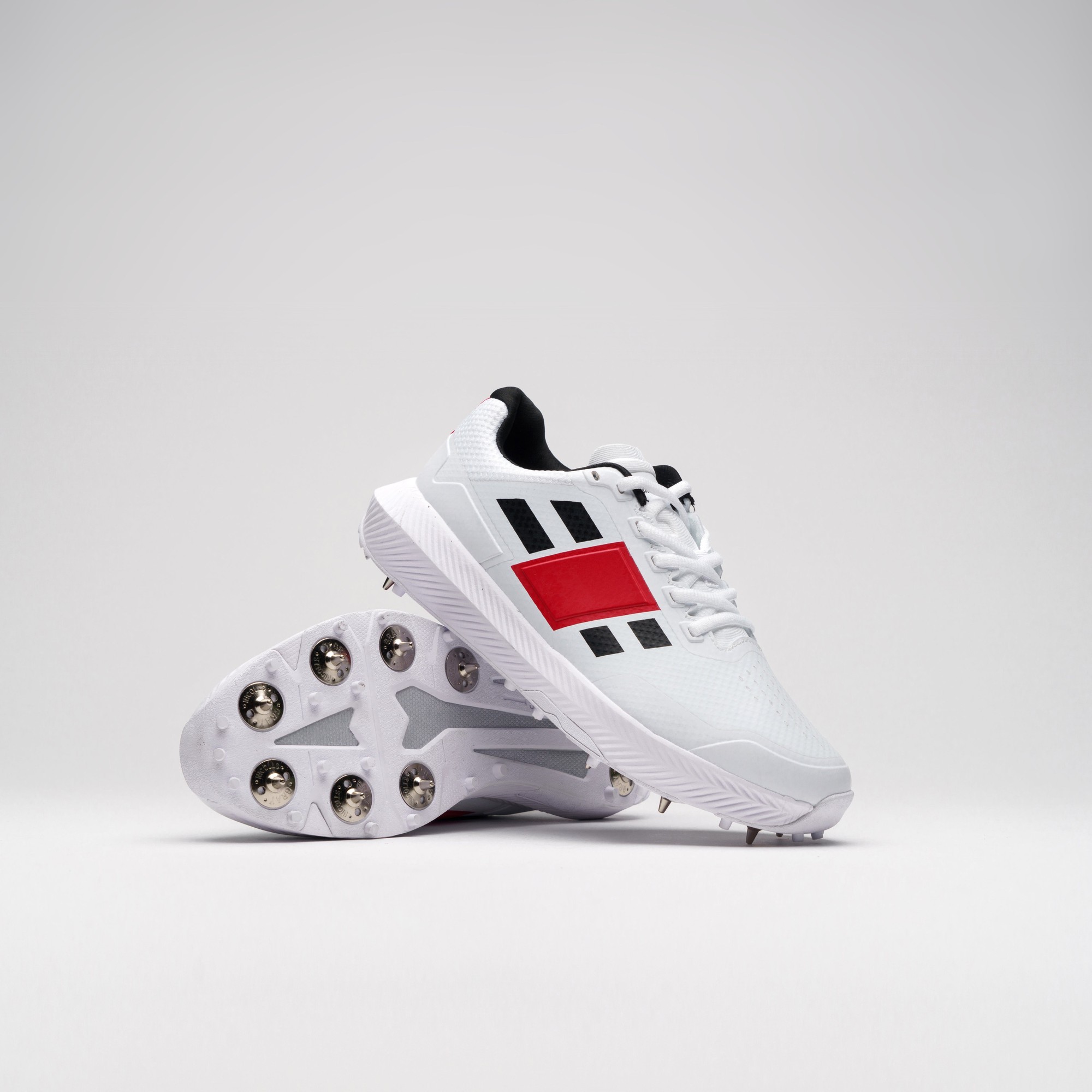 White Gray-Nicolls Revo Pro 1.0 Spike adult shoes feature a breathable mesh upper, black and red accents, and a studded outsole for enhanced traction on the field.