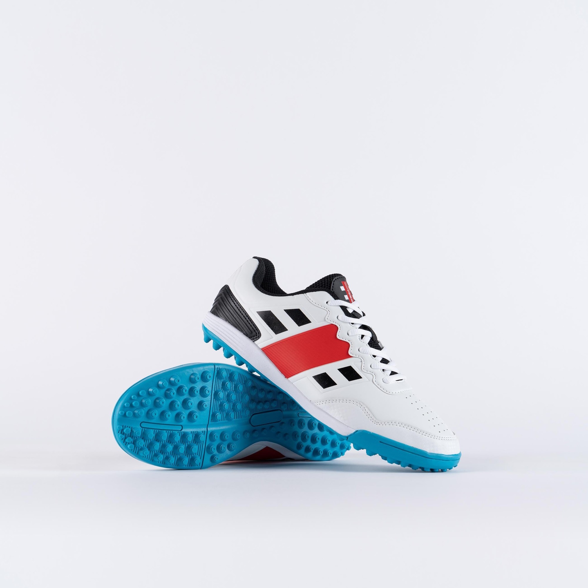 Gray-Nicolls Velocity 3.5 rubber adult shoes feature a white upper with black and red accents, a textured surface, and a blue outsole with prominent studs for traction.