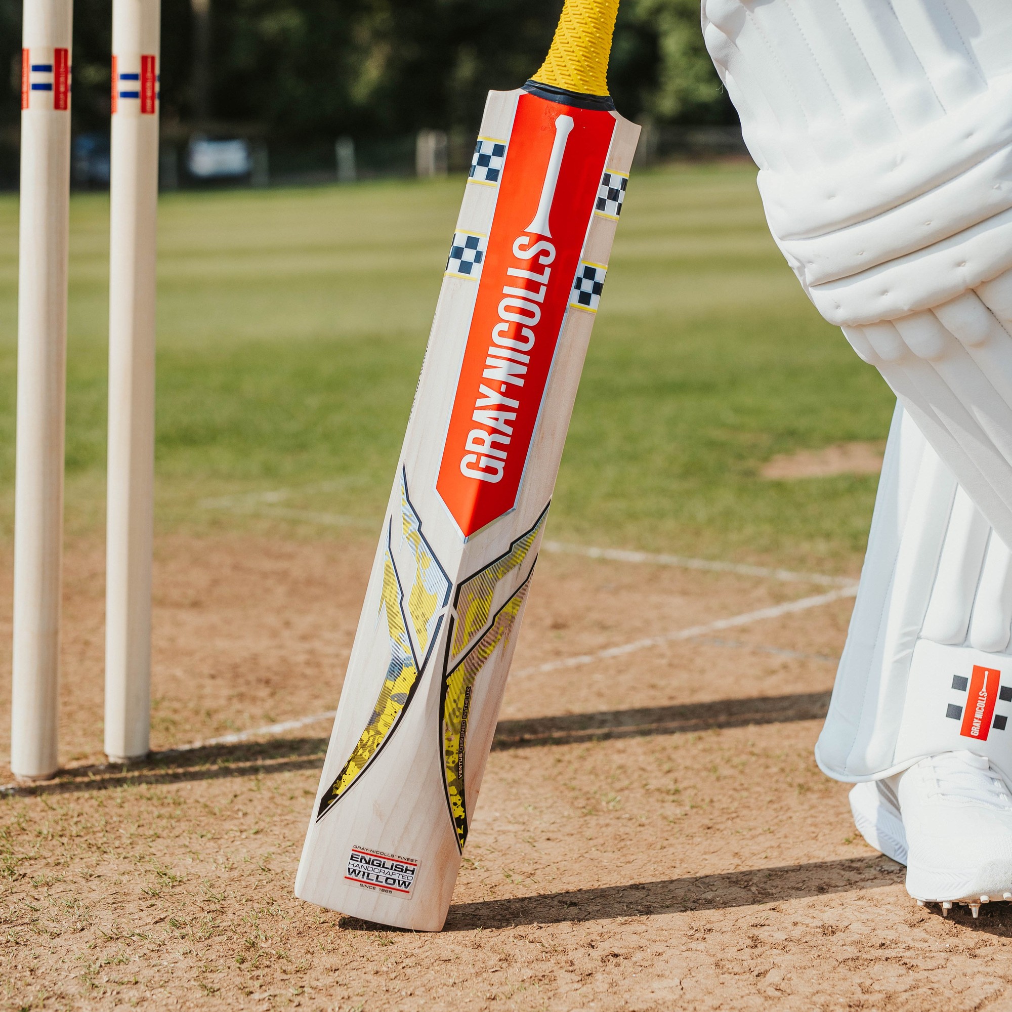 Gray-Nicolls Ventus 1.2 Players Cricket Bat features a sleek design with a white and yellow finish, a prominent red logo, and a yellow grip, resting on the cricket pitch beside stumps.