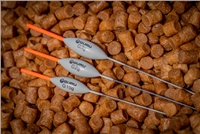 Three Guru F1 Wire Stem Floats in grey with orange tips, featuring a sleek design, resting on a bed of brown fishing pellets, highlighting their lightweight construction.