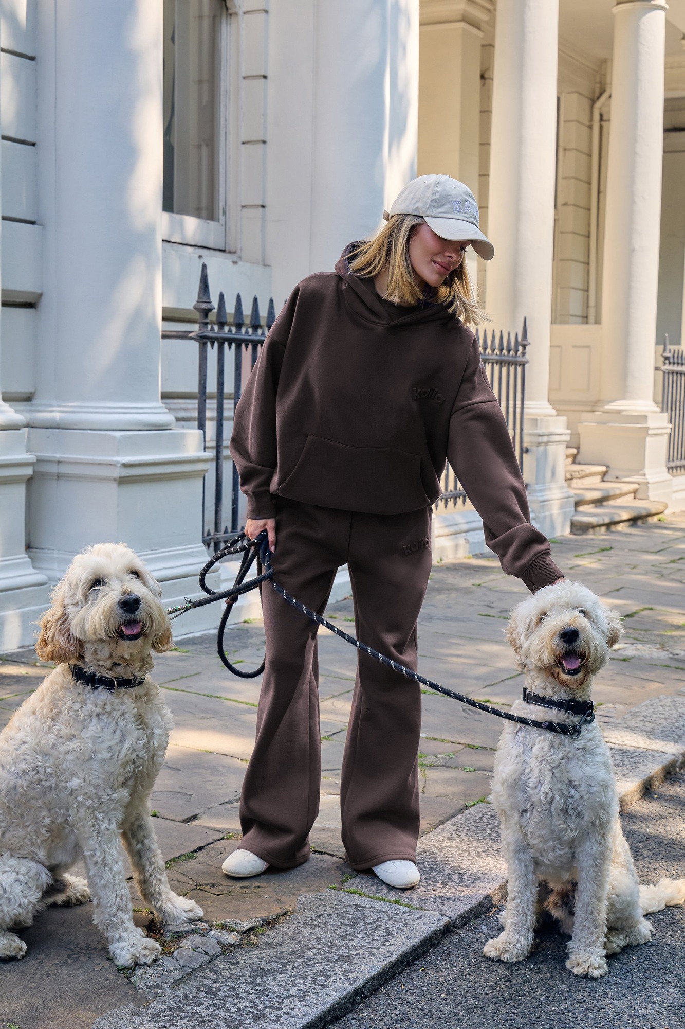 Bitter chocolate wide leg joggers made from embossed fabric, paired with a matching hoodie, worn by a woman walking two fluffy dogs on a city street.
