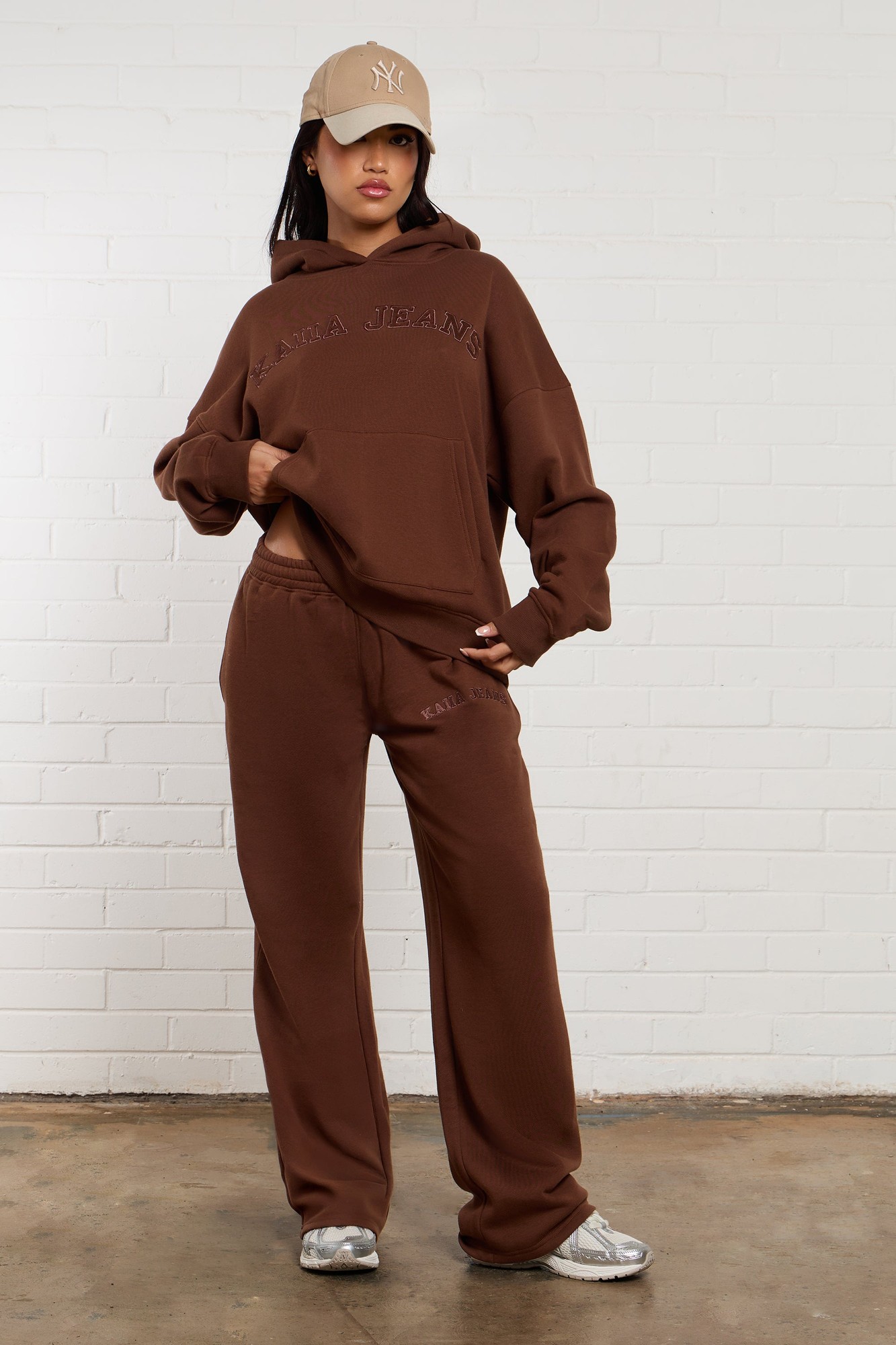 Chestnut-colored wide-leg joggers with embroidered "Kaiia Jeans" branding, paired with a matching oversized hoodie, styled with a beige cap and silver sneakers.