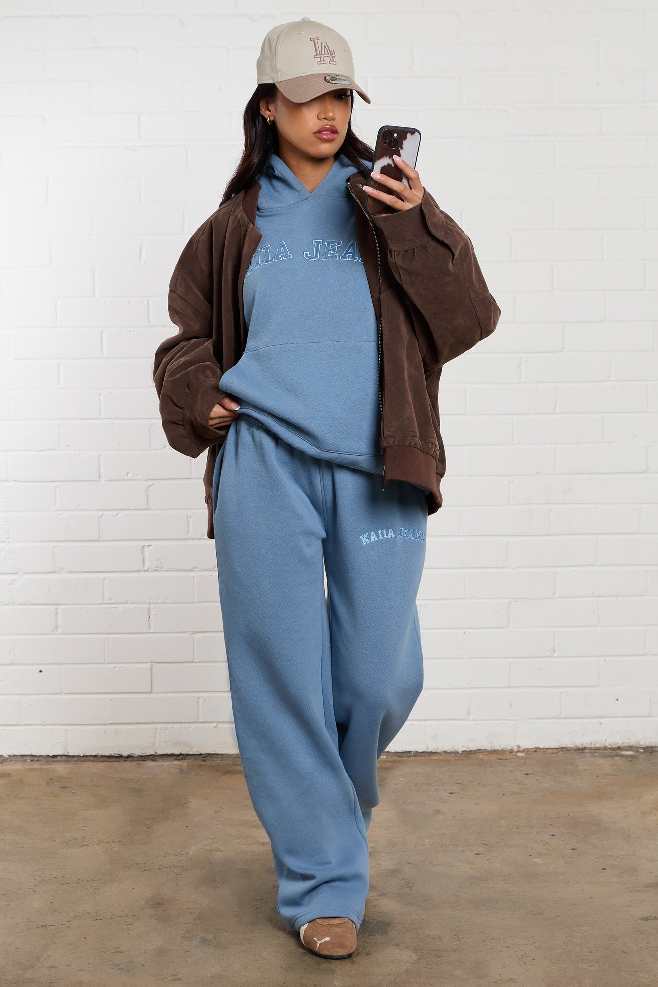 Denim blue wide leg joggers made of soft fabric, featuring embroidered "Kaiia Jeans" logo, styled with a matching hoodie and a brown jacket, complemented by a beige cap.
