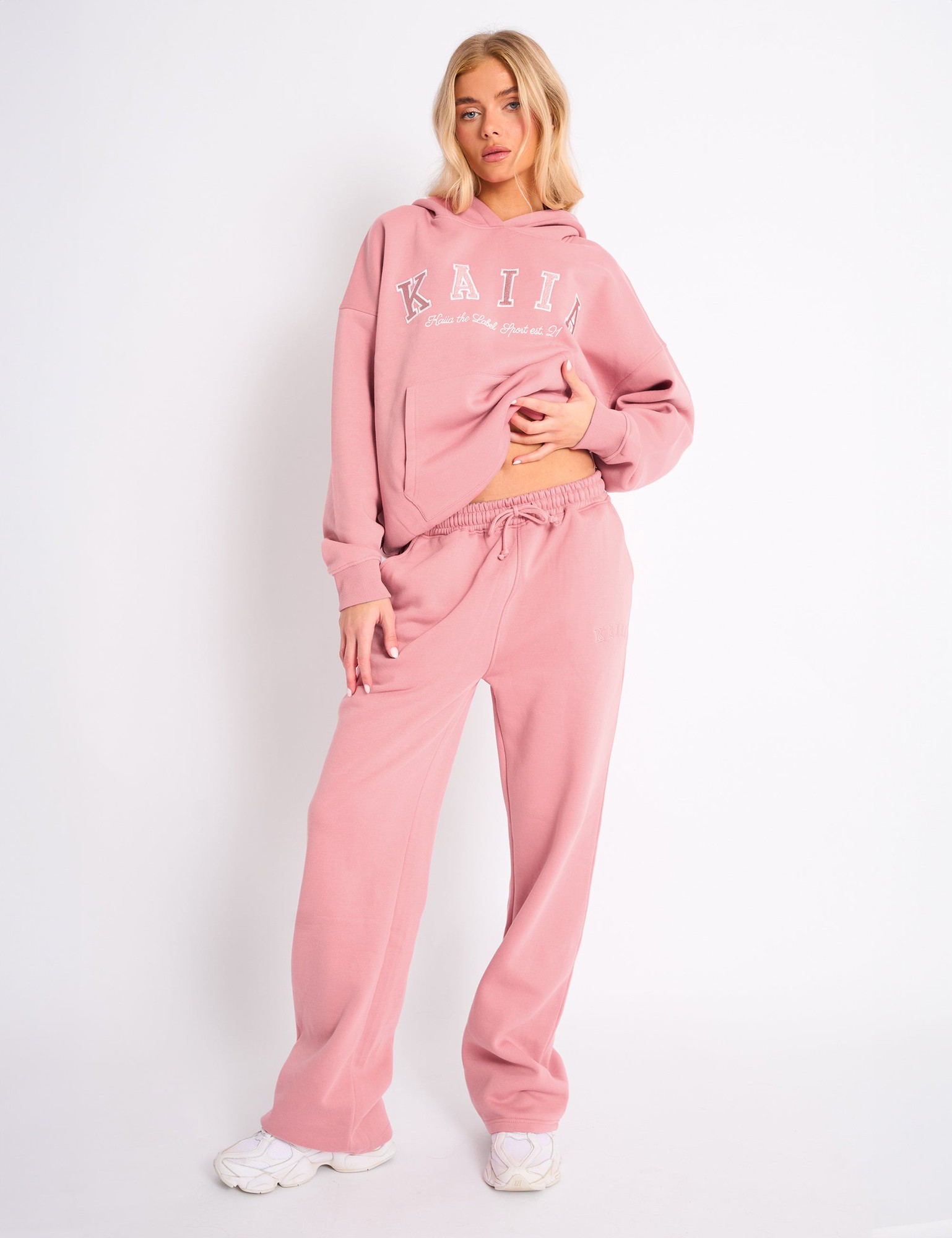 Blush pink wide-leg joggers made of soft material, featuring an elastic waistband with drawstring and subtle logo detailing, styled with a matching hoodie.