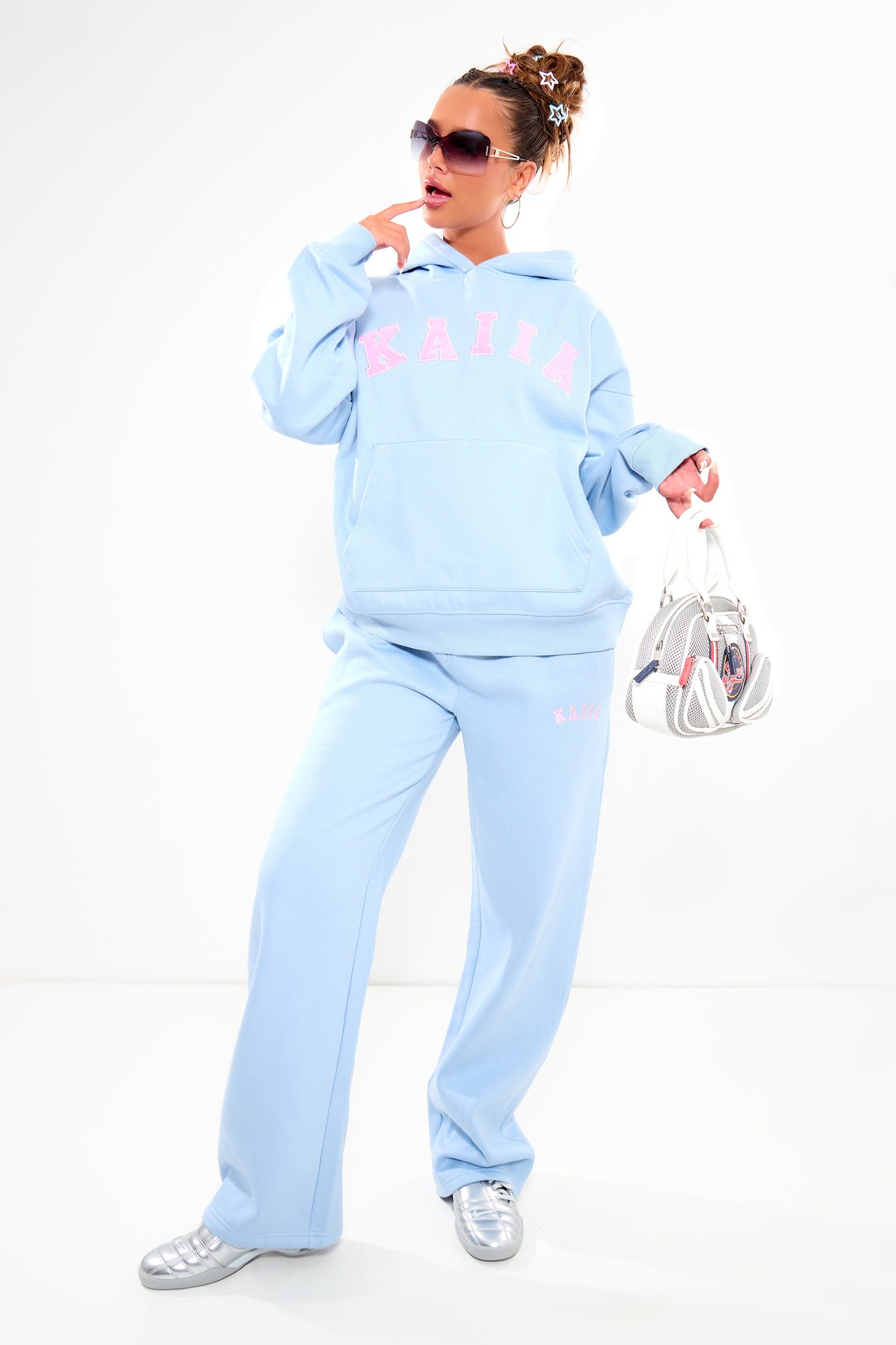 Wide leg joggers in baby blue with pink "KAIIA" logo, made from soft material, paired with a matching hoodie, styled with sunglasses and silver sneakers.