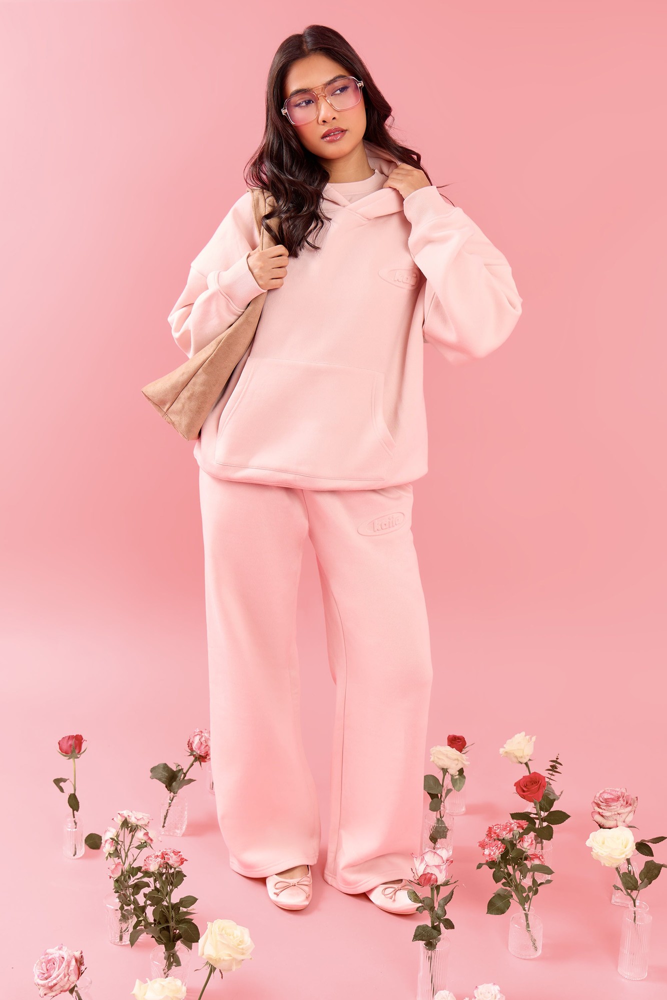 Pink meringue wide-leg joggers with an embossed logo, made from soft material, paired with a matching hoodie and accessorized with a beige bag, surrounded by flowers.