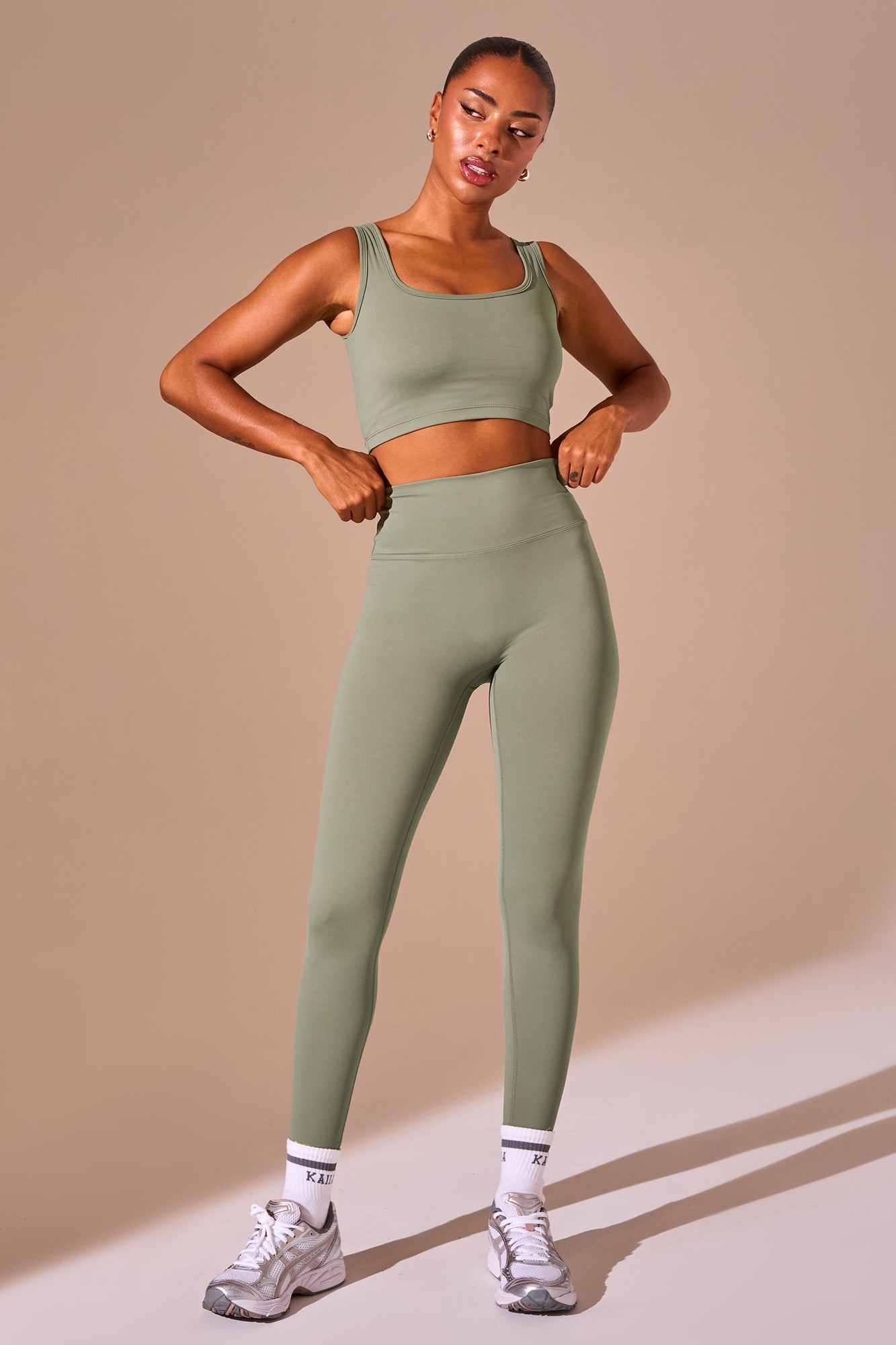 Khaki high-waisted sculpt leggings made from stretchy material, paired with a matching crop top, featuring a fitted silhouette and side cutouts, styled with sporty sneakers.