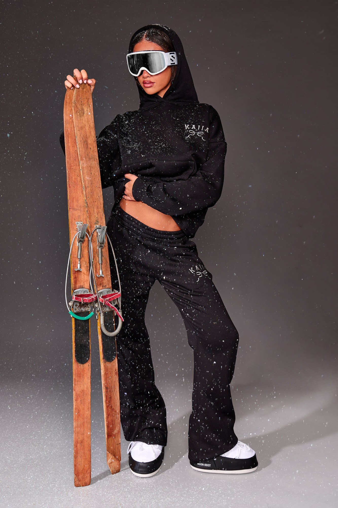 Black flared joggers with silver "Kaiia" slogan, paired with a matching black hoodie, worn by a model holding wooden skis, set against a gray background with snowflakes.