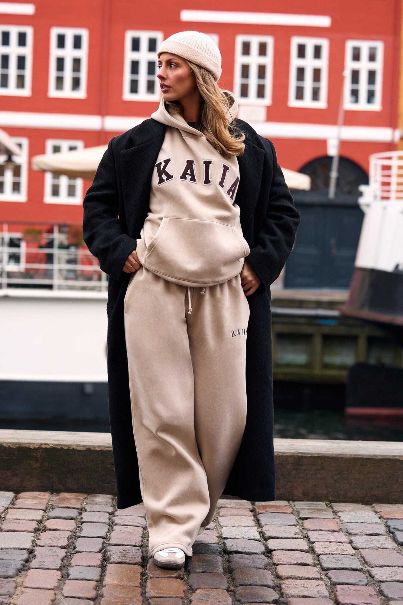 Mid-rise wide-leg joggers in ash taupe with "Kaiia" slogan, paired with a matching hoodie and a black coat, styled with a light beanie and white sneakers.