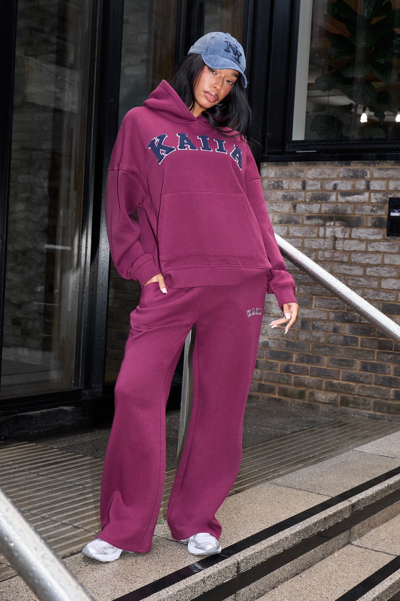 Burgundy wide-leg joggers with a navy logo, paired with a matching burgundy hoodie, worn with a navy cap and white sneakers, set against a brick wall backdrop.