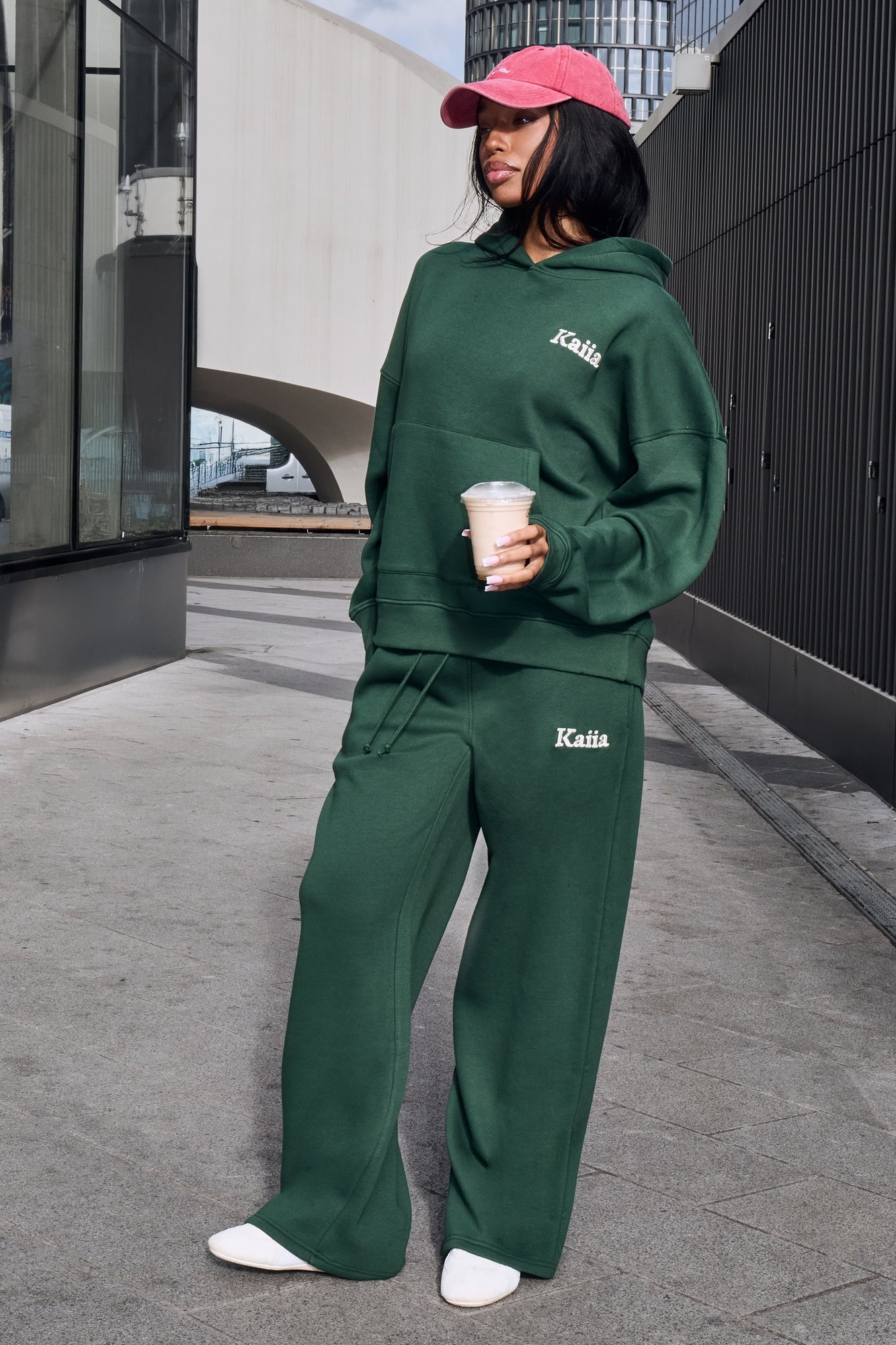 Emerald green wide leg joggers made from soft material, featuring an elastic waistband and the "Kaiia" logo embroidered in white. Paired with a matching hoodie and a pink cap.