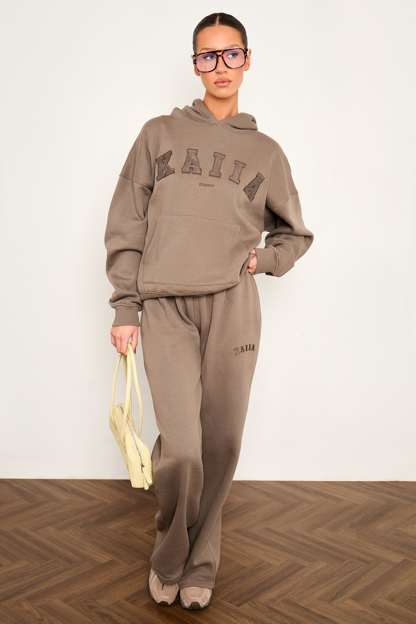 Wide leg joggers in grey taupe featuring the "KAIIA" slogan, made from soft material, styled with a matching hoodie and accessorized with a yellow bag.