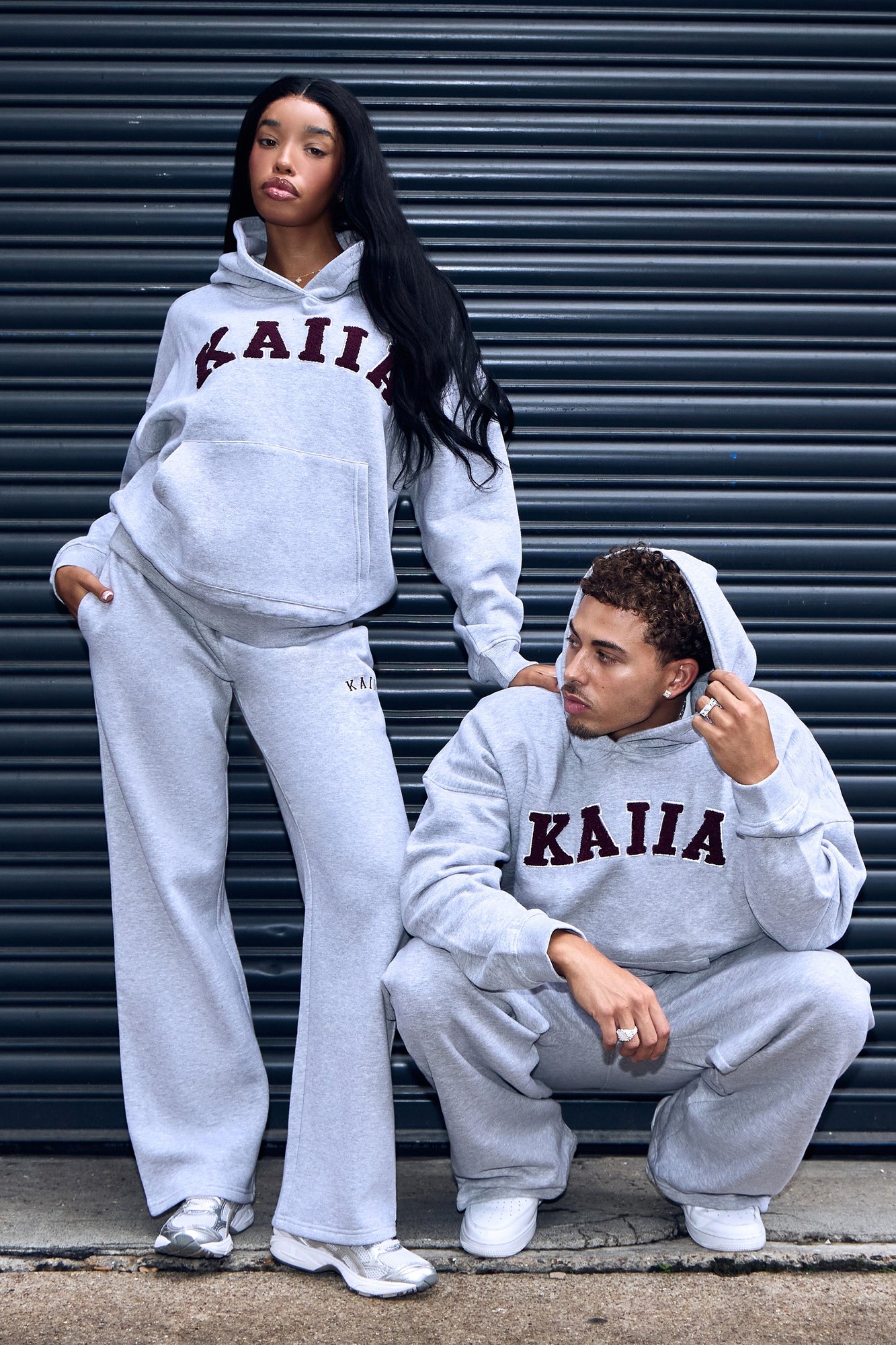 Mid grey wide leg joggers with burgundy "KAIIA" slogan, paired with matching hoodies, showcasing a relaxed fit and soft material, styled by two models against a dark background.