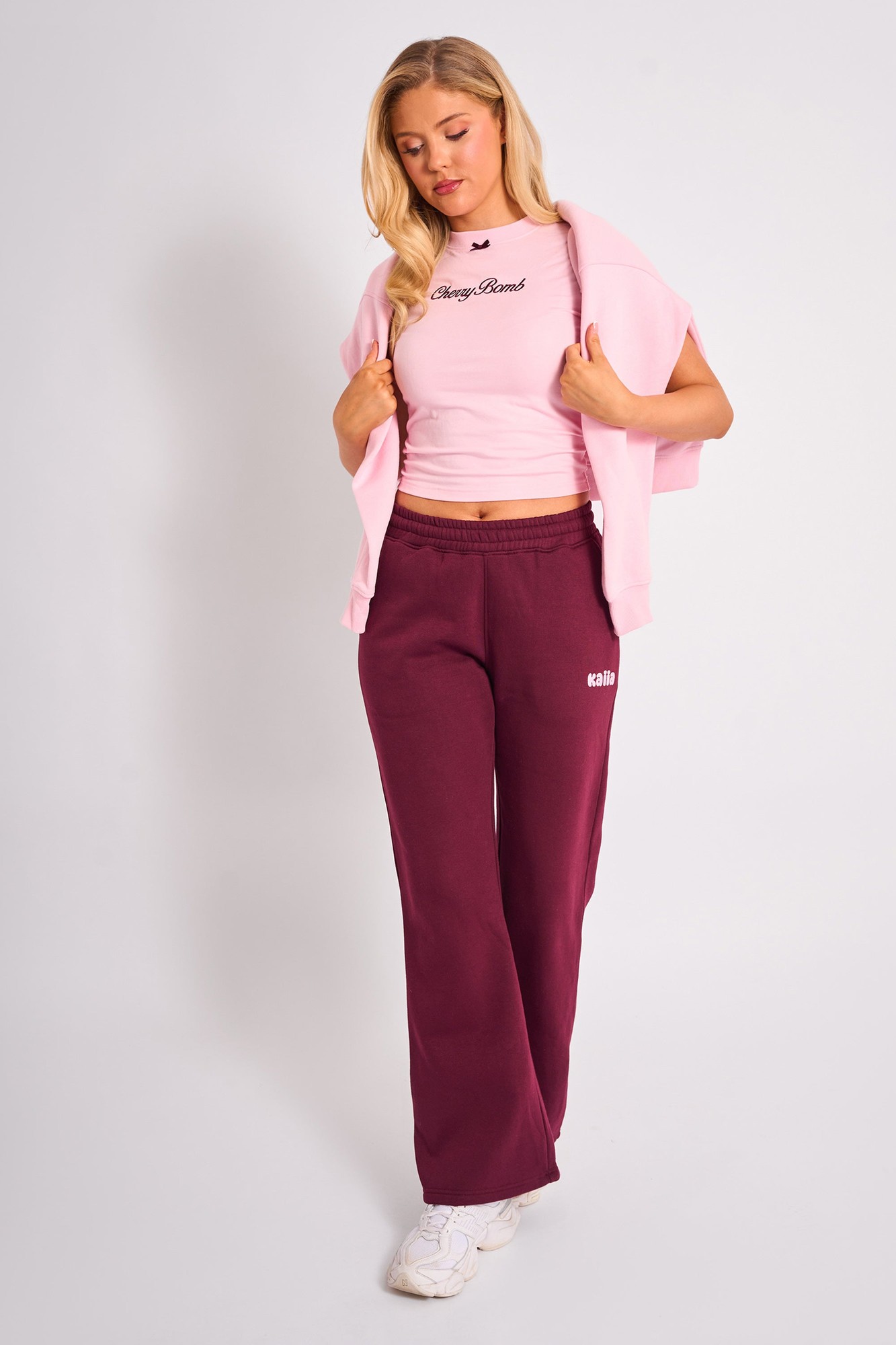 Wide leg joggers in plum with an elastic waistband, paired with a fitted pink crop top, featuring a logo, and a light pink hoodie draped over the shoulders.