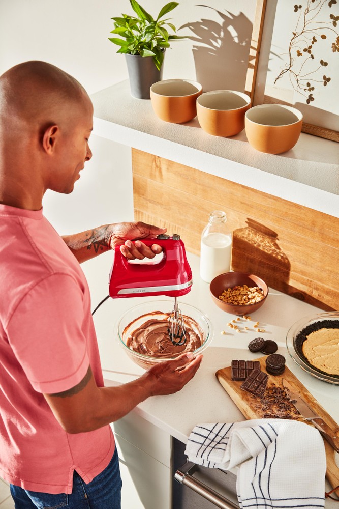 Red KitchenAid hand mixer with two flex edge beaters in use, mixing chocolate batter in a glass bowl, surrounded by ingredients and kitchenware on a countertop.