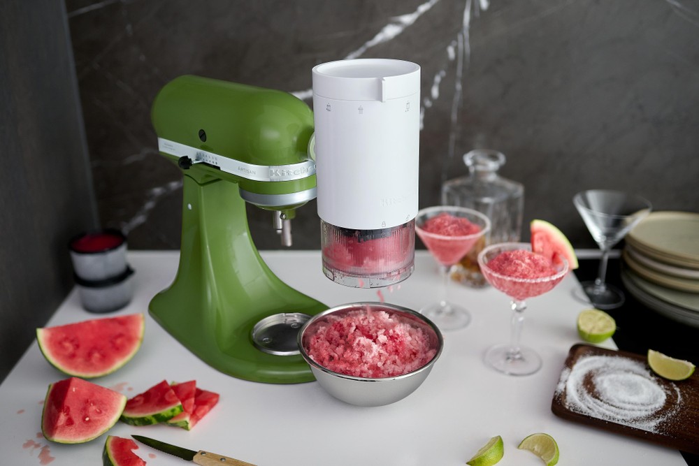 Green KitchenAid stand mixer with a white shaved ice attachment, surrounded by bowls of pink shaved ice, watermelon slices, and cocktails in glasses on a marble countertop.